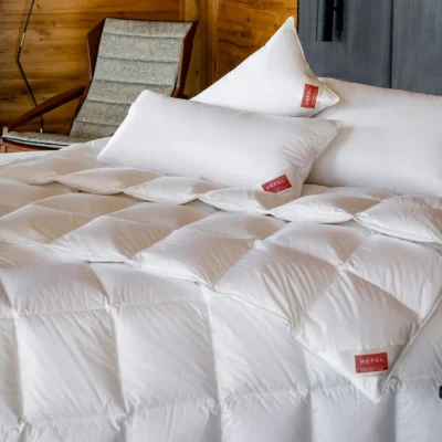 A neatly made bed with a white quilted duvet and several white pillows, all with red labels. A wooden chair with a grey cushion stands in the background against wood-panelled and grey walls.