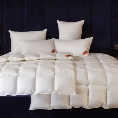 A neatly made bed mit einer whiten, quiltedn duvet und fünf whiten pillow, von denen zwei rote Etiketten haben. Das Bett steht vor einem dunklen, upholstered headboard mit braunen Wandlampen auf jeder Seite.