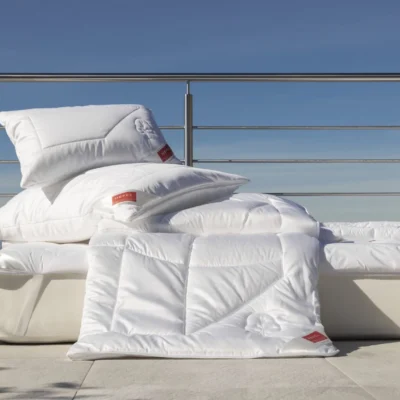 A stack of white pillows and a white duvet from the renowned linen weaving mills, all bearing red labels, neatly placed on a white outdoor lounge chair against a backdrop of clear blue sky and metal railings.
