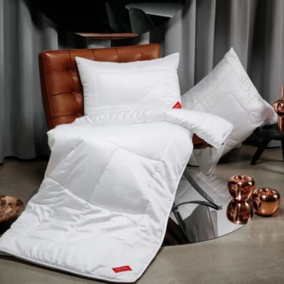 A modern room with a light brown leather armchair draped with a white duvet and a pillow, plus another pillow on a black stool. Copper decorative items and grey curtains complete the modern ambience.