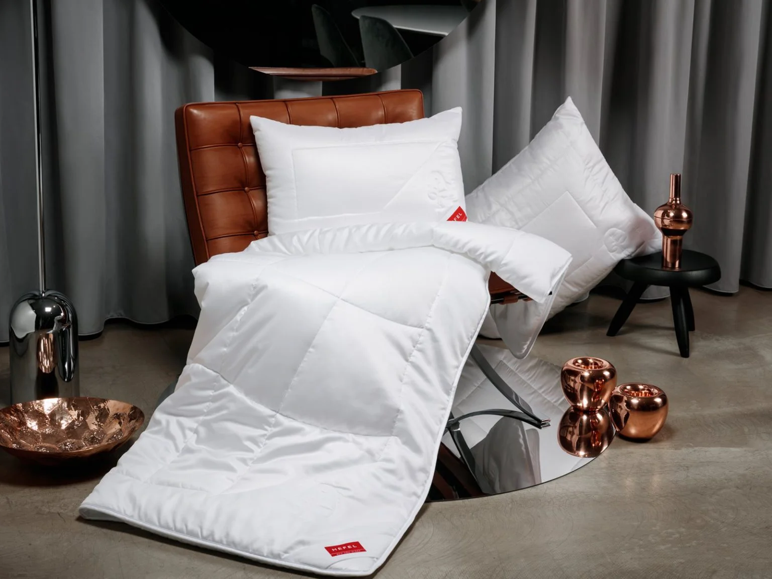 Ein modernes Zimmer mit einem hellbraunen Ledersessel, der mit einer weißen Decke und einem Kissen drapiert ist, sowie einem weiteren Kissen auf einem schwarzen Hocker. Kupferne Dekoartikel und graue Vorhänge vervollständigen das moderne Ambiente.