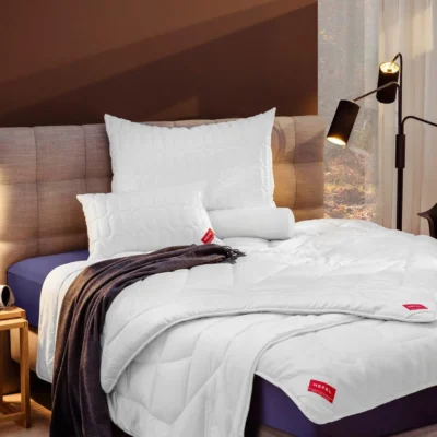 A neatly made bed with an upholstered headboard, white bedding and pillows, a dark throw, two modern black floor lamps and a window with sheer curtains letting in natural light.