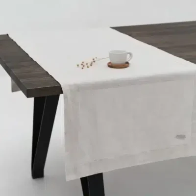 Ein dunkler Holztisch mit schwarzen Beinen ist mit einem weißen Leinentischläufer aus renommierten Leinenwebereien bedeckt. Auf dem Läufer stehen eine weiße Tasse und Untertasse sowie eine kleine getrocknete Pflanze. Der Hintergrund ist weiß und minimalistisch.
