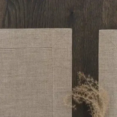 Zwei beigefarbene Stoff-Tischsets liegen auf einer dunklen Holzfläche, wobei sich die Ecken fast berühren. Ein kleiner Zweig mit getrocknetem Gras liegt teilweise auf dem rechten Tischset.