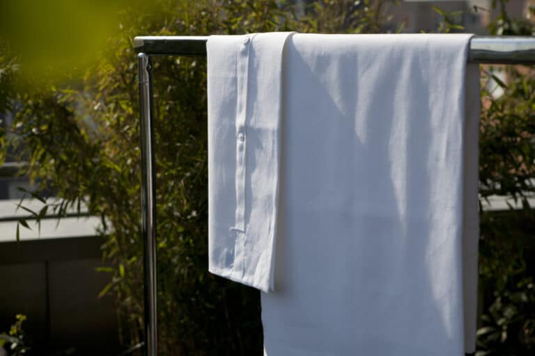 A white sheet and pillowcase hang over a metal clothes rack outside, with green leafy plants in the background. The sunlight shines on the fabric and emphasizes its clean appearance.