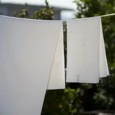 Zwei weiße Hemden, die auf einer Wäscheleine im Freien hängen und im Sonnenlicht trocknen, mit grünen Bäumen und einem unscharfen Gebäude im Hintergrund.