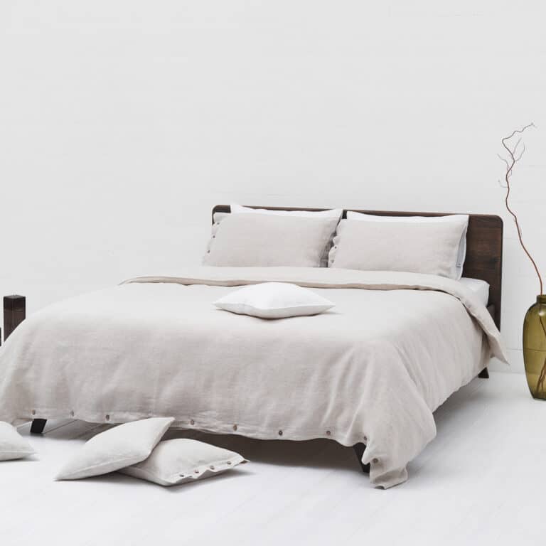 A modern bed with beige linen bedding and pillows stands in front of a white wall. Two pillows lie on the bed, three more are scattered on the white floor, and nearby stands a large green vase with a dry branch.