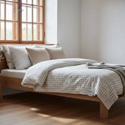 Ein ordentlich gemachtes Bett mit beige-weiß kariertem Bettzeug, zwei beigefarbenen Kissen und einer grauen Decke in einem hellen Zimmer mit Holzfußboden und einem großen Fenster, das natürliches Licht hereinlässt.