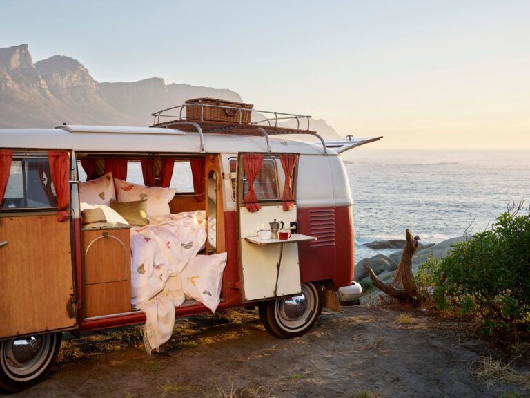 An old red and white caravan, as pictured in Linen Magazine, is parked on a cliff by the sea. Its door opens to a cozy bed with a picnic basket on the roof and mountains rising under a soft golden sky in the background.