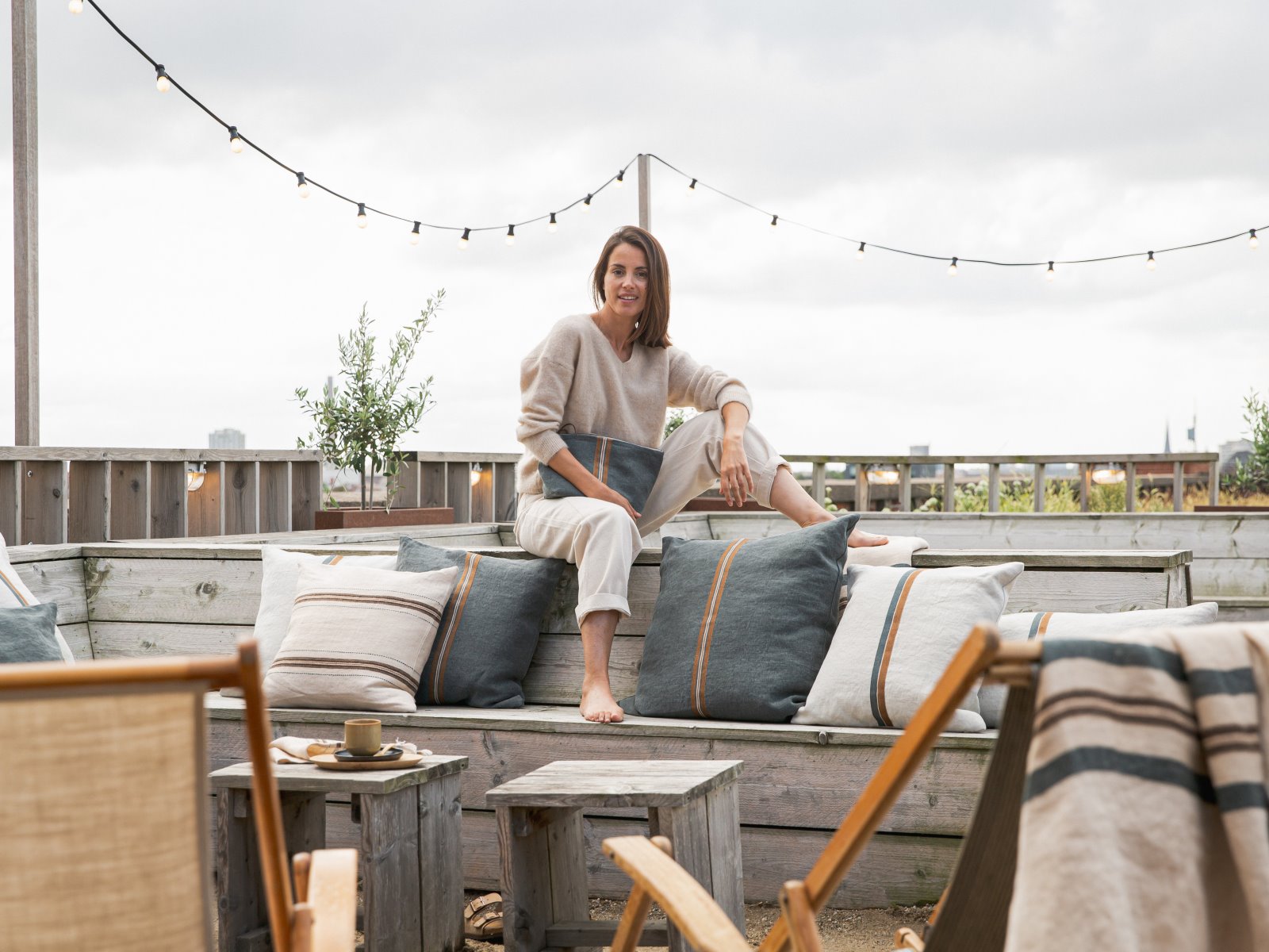 A woman sits barefoot on an outdoor wooden bench with cushions and smiles at the camera. String lights hang above, and patio chairs and a coffee cup are visible in the cozy, relaxed setting.
