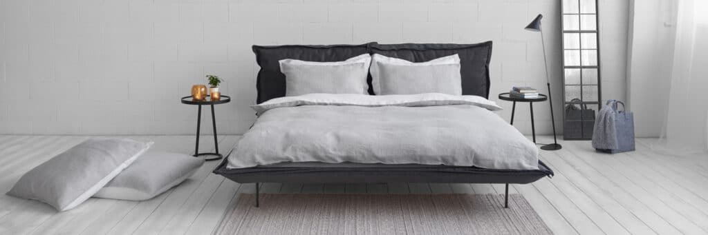 Ein modernes Schlafzimmer hat ein ordentlich gemachtes Bett mit grauer Bettwäsche, zwei Kissen auf dem Boden, schwarze Beistelltische mit Dekor, eine hohe Stehlampe, einen Standspiegel und eine graue Tasche auf hellem Holzboden.