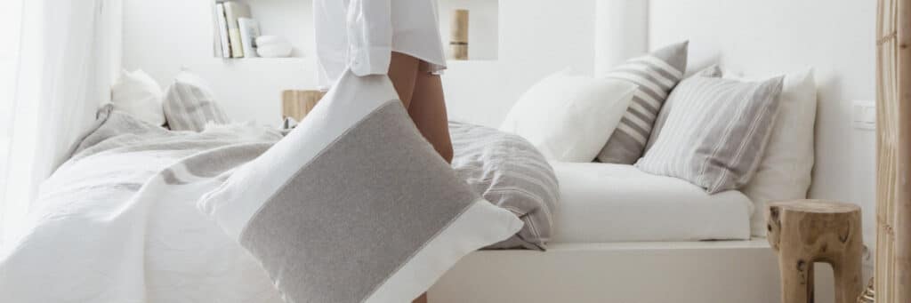 Eine Person steht neben einem ordentlich gemachten Bett in einem hellen, minimalistischen Schlafzimmer und hält ein graues und weißes Kissen. Der Raum ist in neutralen Tönen gehalten, mit weicher Bettwäsche und schlichtem Dekor.