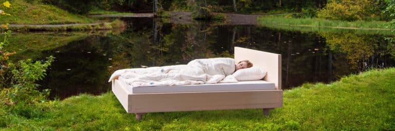 A person sleeps in a bed with white bedding on a lawn outdoors near a tranquil pond, surrounded by green trees and leaves.
