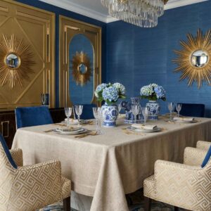 Elegantes Esszimmer mit Salome Reinleinen-Tischdecken von Schlitzer Leinen, blauem und goldenem Dekor, blauen Hortensien in gemusterten Vasen, goldenen Wandspiegeln mit Sonnenschliff, blauen Wänden, gemusterten Stühlen und einem auffälligen Kristalllüster.