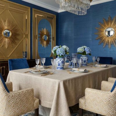 Elegant dining room with Salome Pure Linen Tablecloths by Schlitzer Leinen, blue and gold decor, blue hydrangeas in patterned vases, gold sunburst wall mirrors, blue walls, patterned chairs and a striking crystal chandelier.