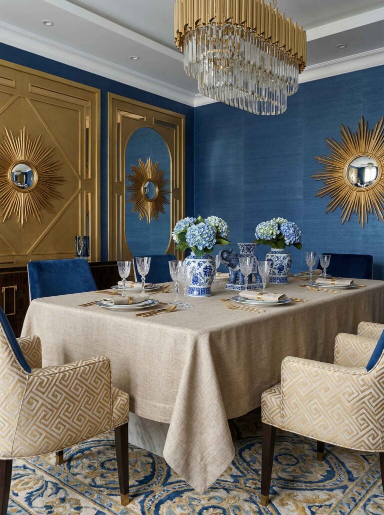 Elegantes Esszimmer mit Salome Reinleinen-Tischdecken von Schlitzer Leinen, blauem und goldenem Dekor, blauen Hortensien in gemusterten Vasen, goldenen Wandspiegeln mit Sonnenschliff, blauen Wänden, gemusterten Stühlen und einem auffälligen Kristalllüster.