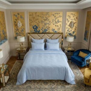 Elegantes Schlafzimmer mit goldener und blauer Blumentapete, einem Bett mit Sol Halbleinen-Bettwäsche von Schlitzer Leinen, verspiegelten Nachttischen, zwei Lampen und einem goldenen Kopfteil. Blaue und gelbe Stühle stehen an einem großen Fenster mit durchsichtigen Vorhängen.