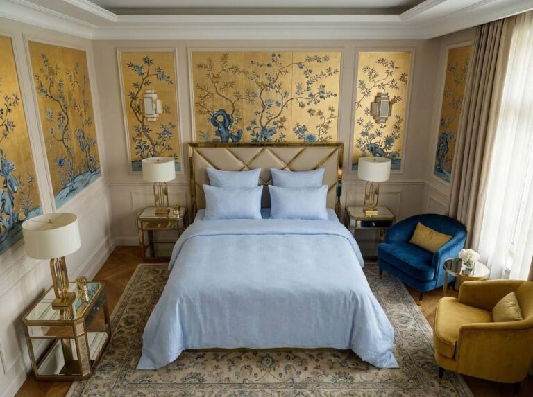 Elegant bedroom with gold and blue floral wallpaper, a bed with Sol half-linen bedding by Schlitzer Leinen, mirrored bedside tables, two lamps and a gold headboard. Blue and yellow chairs stand by a large window with sheer curtains.