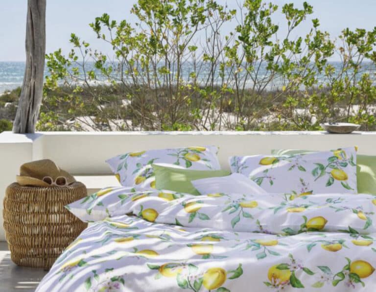 Bett im Freien mit zitronengemusterter Bettwäsche aus dem Linen Magazine, neben einem Pool und im Schatten einer Holzpergola. In der Nähe steht ein Weidenkorb mit Hut und Sonnenbrille. Der Sandstrand, das Grün und das Meer glitzern unter einem klaren Himmel.