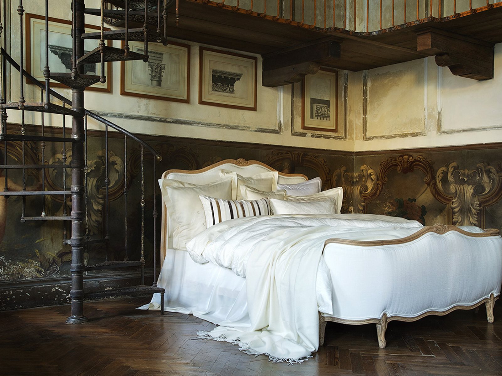 A luxurious bed with white linens stands in an ornate vintage-style bedroom—worthy of a feature in Linen Magazine—with carved wood decor, framed art on the walls, and a wrought-iron spiral staircase nearby.
