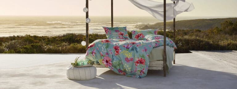Ein Himmelbett mit lebhaft geblümter Bettwäsche steht auf einer glatten Terrasse in der Nähe von Grünpflanzen und bietet einen Blick auf das Meer bei Sonnenuntergang. Weiße Vorhänge hängen an den Holzpfosten, und ein kleiner runder Hocker mit einer Pflanze steht in der Nähe.