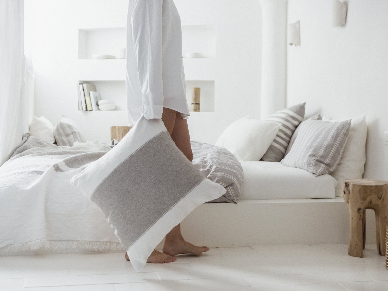 Eine Person in einem weißen Hemd hält ein grau-weißes Charlotte Leinen Plaid von Libeco-Kissen und steht barfuß neben einem ordentlich gemachten Bett in einem hellen, minimalistischen Schlafzimmer mit neutralen Tönen und sanfter Beleuchtung.