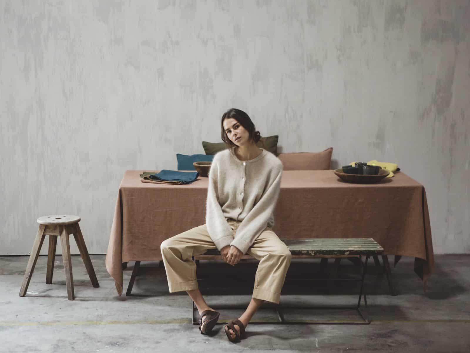 Eine Frau in einem gemütlichen Pullover und einer beigen Hose sitzt an einem mit Hudson Optic White - reines Leinen für den Tisch von Libeco geschmückten Esstisch in einem minimalistischen, neutral getönten Raum mit Betonböden und einer einfachen Kulisse.