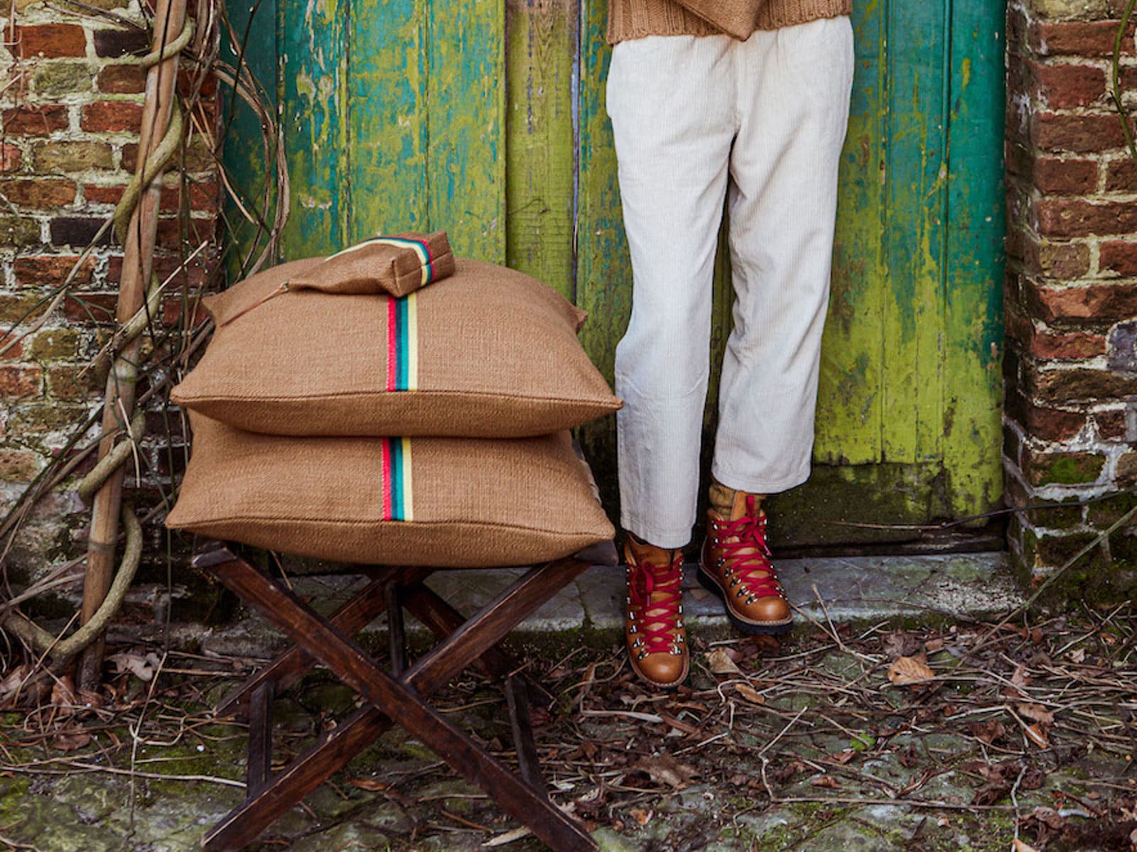 Eine Person in hellen Hosen und roten Schnürstiefeln steht vor einer verwitterten grünen Tür. Daneben steht ein Holzschemel mit zwei gestapelten Jutekissen, die beide mit einem bunten Streifen versehen sind. Getrocknete Blätter bedecken den Boden.