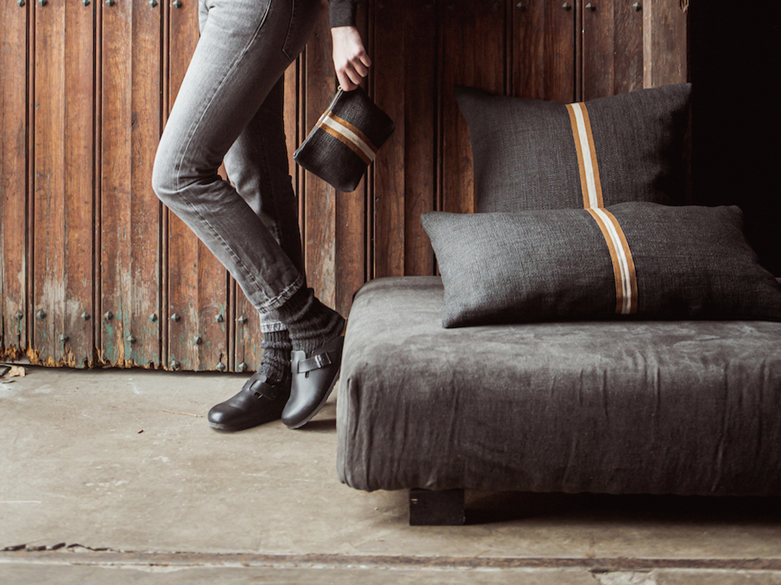 Eine Person in grauen Jeans und schwarzen Schuhen lehnt an einer Holzwand und hält eine kleine dunkle Stofftasche. Zwei dunkle Kissen mit hellbraunen Streifen liegen auf einem passenden niedrigen Sofa in der Nähe.