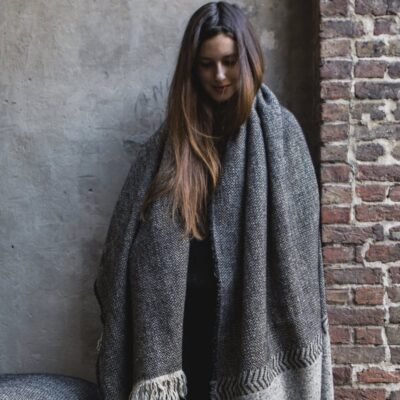Eine Frau mit langen braunen Haaren steht vor einer strukturierten Wand, eingewickelt in die Decke Jules Black Herringbone Leinen-Wolle-Mix von Libeco, und blickt mit sanftem Blick nach unten.