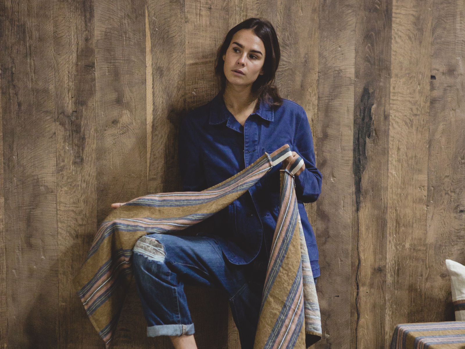 Eine Frau in blauem Jeanshemd und Jeans steht an einer Holzwand, hält das Olympia - reines Leinenhandtuch von Libeco in der Hand und blickt nachdenklich zur Seite.