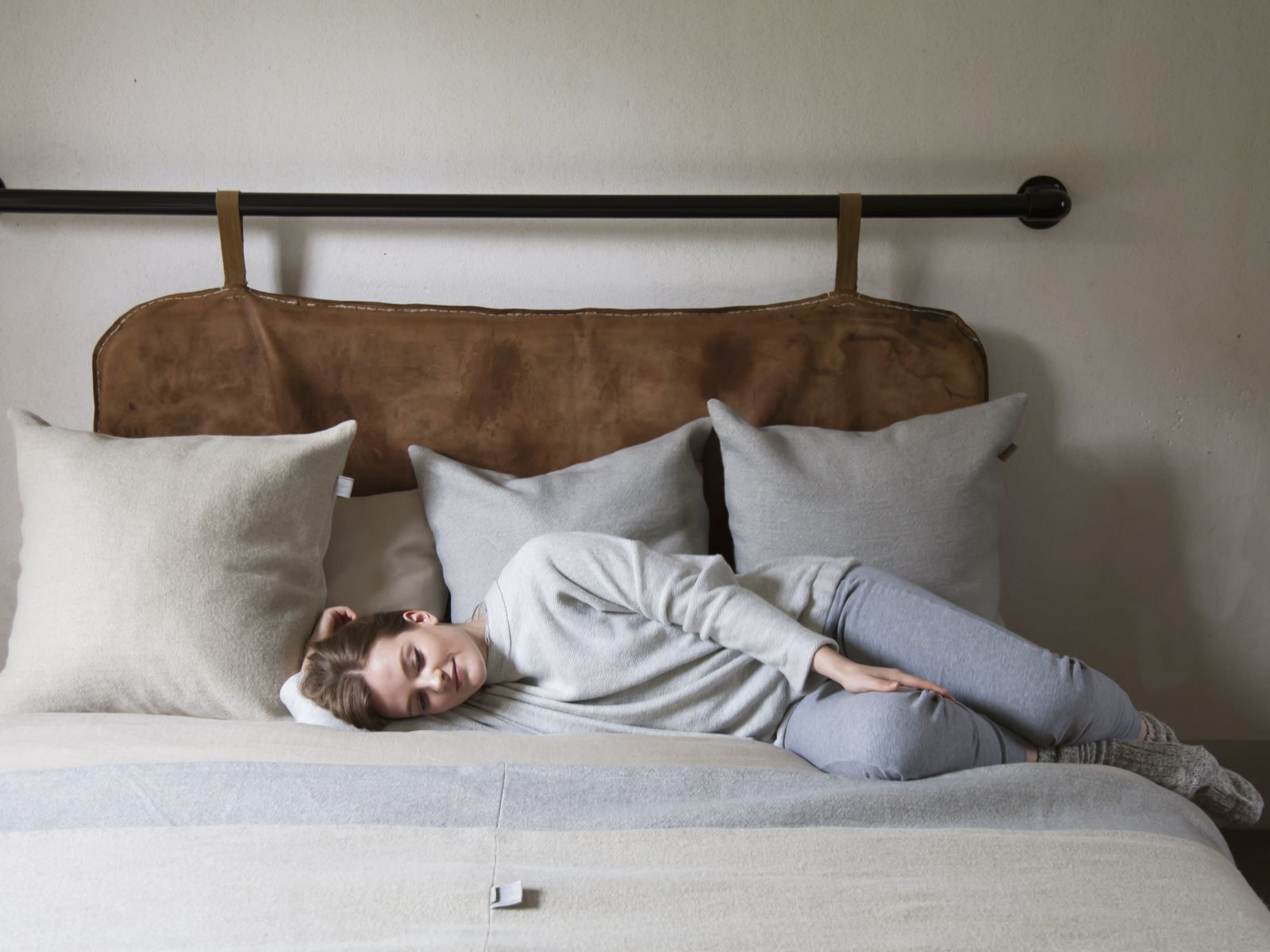 Eine Person mit kurzen Haaren in einem grauen Outfit liegt auf der Seite auf einem Bett mit beigen und grauen Kissen und stützt ihren Kopf auf ein Kissen in einem gemütlichen, minimalistischen Schlafzimmer.