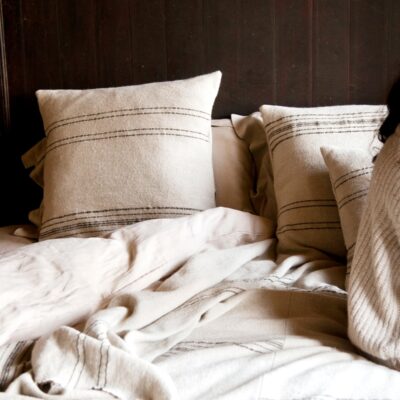 Auf einem gemütlichen Bett liegt der Marokkanische Streifen Leinen-Wolle-Mix von Libeco in beige-weiß gestreift. Auf der rechten Seite sitzt eine Person mit langen dunklen Haaren in einem hellen Strickpullover, der eine warme, entspannte Atmosphäre schafft.