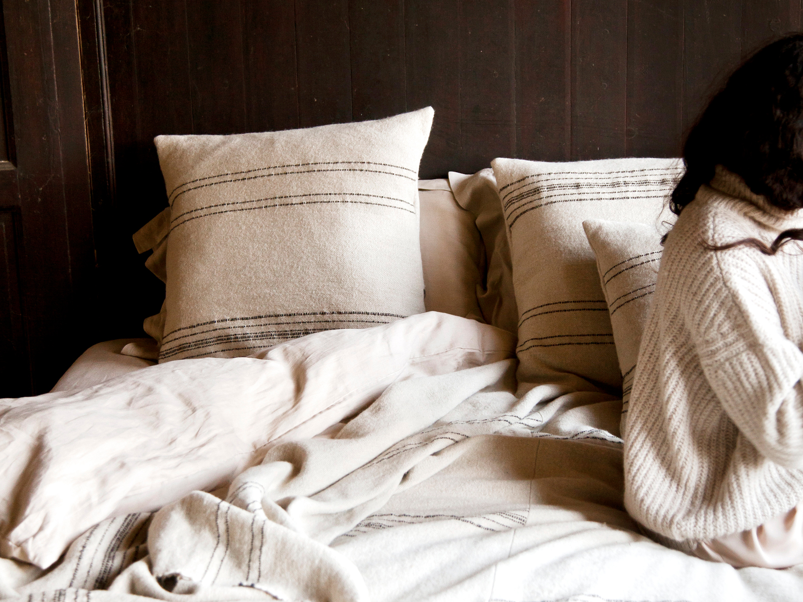 Auf einem gemütlichen Bett liegt der Marokkanische Streifen Leinen-Wolle-Mix von Libeco in beige-weiß gestreift. Auf der rechten Seite sitzt eine Person mit langen dunklen Haaren in einem hellen Strickpullover, der eine warme, entspannte Atmosphäre schafft.
