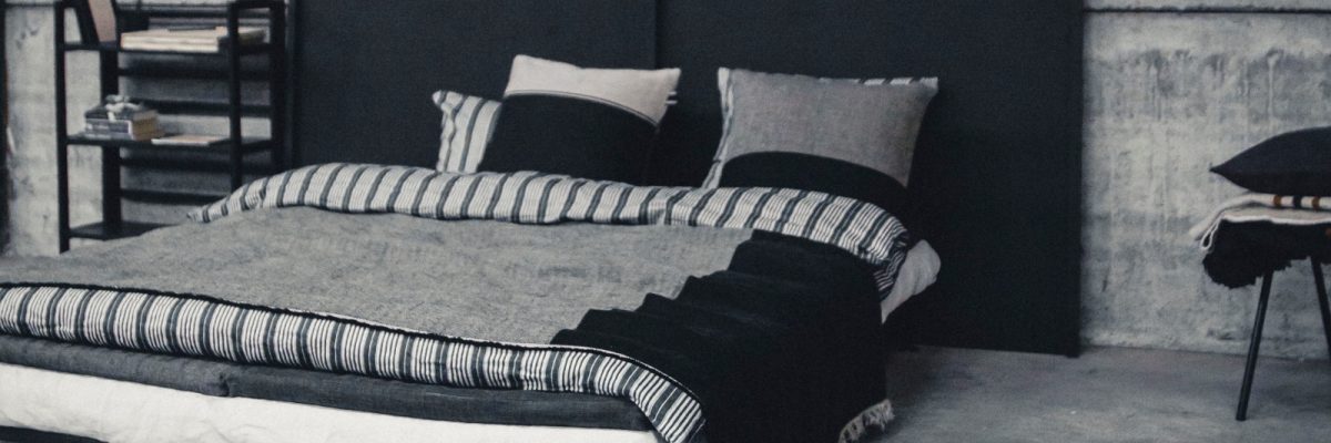 Ein modernes Schlafzimmer mit einem niedrigen Bett mit einfarbig gestreiftem Bettzeug und Kissen vor einem dunklen Kopfteil und einer Betonwand. Ein kleines Regal mit Büchern und ein schwarzer Stuhl mit gefalteten Decken stehen daneben.