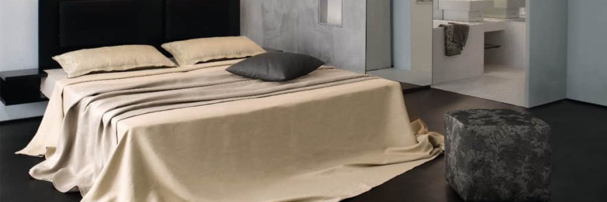 Ein modernes Schlafzimmer mit einem großen Bett, das mit beiger Uni Stoffe - einzig und allein Bettwäsche, zwei Kissen und einem grauen Kissen bezogen ist. In der Nähe steht ein grauer Hocker mit Textur, der den Blick auf ein schlichtes Badezimmer mit weißem Waschbecken und Spiegel freigibt.
