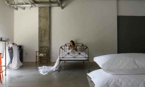 Eine Frau sitzt auf einem Metallbett in einem minimalistischen Raum im Industriestil mit Betonboden und freiliegenden Rohren. Große weiße Kissen sind im Vordergrund gestapelt. Von links strömt Licht herein.