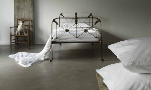 Ein metallgerahmtes Bett mit weißer Bettwäsche steht in einem minimalistischen Raum mit Betonboden. Neben dem Bett steht ein Stuhl mit einem kleinen Hund darauf, und weiße Laken ziehen sich vom Bett bis zum Boden. Zwei Kissen befinden sich im Vordergrund.