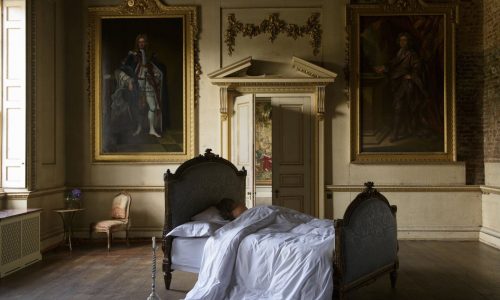 Eine Person schläft in einem antiken Bett, gekleidet in Sea Island Bettwäsche von Peter Reed, umgeben von gerahmten Porträts, eleganten Stuckarbeiten, hohen Decken und großen Fenstern, die Licht in den großen klassischen Raum werfen.