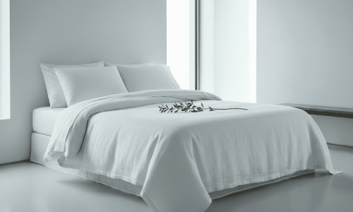 Ein modernes, minimalistisches Schlafzimmer mit Leinen Bettwäsche Fischgrat in Weiß von Vieböck 273, zwei Kissen, einem Ast mit grünen Blättern auf dem Bett, großen Fenstern und einer einfachen Holzbank vor einer schlichten weißen Wand.