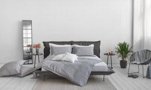 Modern bedroom with a grey bed in Arctica Classic Linen Bedding by Geniksa, white and grey bedding, pillows on the floor, a side table with candles and a plant, a chair and a tall mirror against a white wall and light wooden floor.
