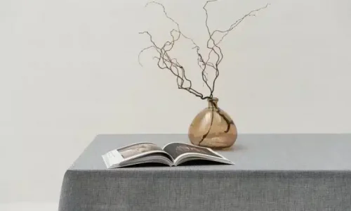 Eine einfache Szene mit einem mit grauer Tischdecke bedeckten Tisch, auf dem ein aufgeschlagenes Buch liegt, und einer braunen Glasvase mit kahlen, sich windenden Zweigen vor einem schlichten hellen Hintergrund.