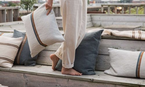Eine leicht bekleidete Person steht barfuß auf einer hölzernen Sitzgelegenheit im Freien und hält ein Kissen von Atlas Natural reines Leinen von Libeco in der Hand; passende Kissen sind in der Nähe angeordnet, Pflanzen und eine Stadtkulisse sind zu sehen.