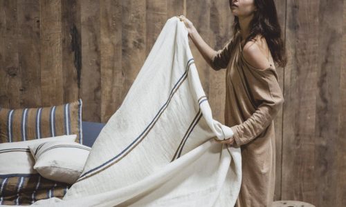 Eine Frau in einem lockeren, hellbraunen Kleid macht ein Bett mit cremefarbener und blau gestreifter Bettwäsche mit dem Auburn Leinen Coverlet und Kissen von Libeco in einem rustikalen Raum mit Holzwänden und gestreiften Kissen.