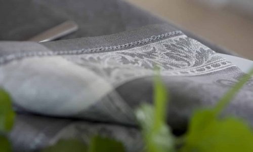 Nahaufnahme eines gefalteten grauen Stoffes mit einem verschlungenen weißen Blumenmuster, der auf einer Fläche mit grünen Blättern im Vordergrund ruht.