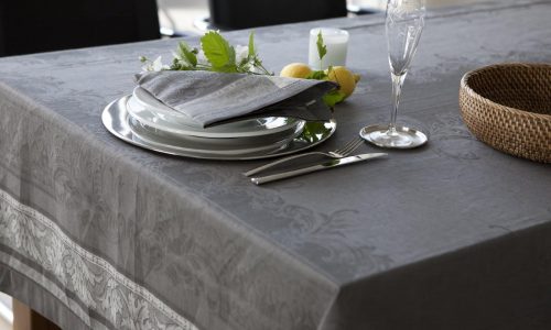 Auf einem Esstisch stehen die Tischdecke aus Leinen Damast Chrysantheme von Hoffmann, gestapelte weiße Teller, eine graue gefaltete Serviette, Silberbesteck, Glas, Kerze, ein geflochtener Korb und ein kleines Arrangement aus grünen Blättern mit Zitronen.