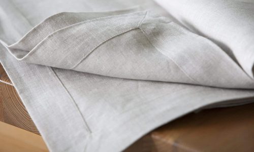 Eine Nahaufnahme einer hellgrauen Leinentischdecke, die ordentlich über einen Holztisch drapiert ist, mit sauberen Nähten und einer strukturierten Stoffoberfläche.