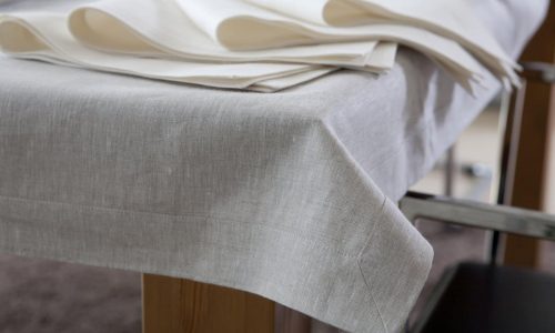 Nahaufnahme einer beigen Leinentischdecke, die einen Holztisch bedeckt, mit ordentlich gefalteten cremefarbenen Servietten, die am Rand platziert sind. Ein Metallstuhl ist teilweise neben dem Tisch zu sehen.