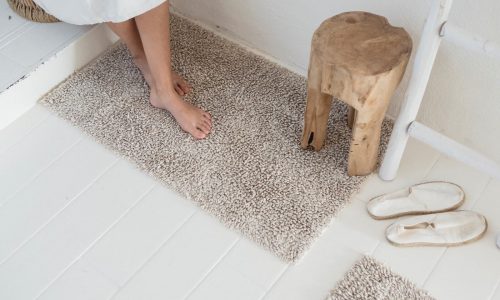 Eine weiß gekleidete Person sitzt barfuß auf einer weißen Stufe neben einem kleinen Holzschemel. Auf dem weißen Boden liegen weiße Hausschuhe und ein Calistoga Leinenteppich fürs Bad von Libeco.