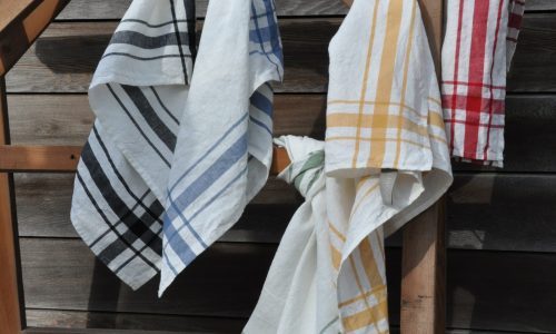 Das Camaret Black - reines Leinen für die Küche von Libeco und andere gestreifte Küchentücher in Schwarz, Blau, Gelb, Grün und Rot hängen in einem Holzrahmen an einer verwitterten Holzwand.